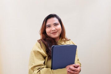 Fototapeta premium Warm smile radiates from a confident woman holding a clipboard. Her hands grip the folder, poised for work or presentation. Soft beige backdrop highlights her calm, professional demeanor.