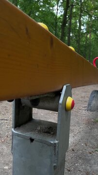 Low angle seesaw person silhouette background, wooden plank leading to red handle and yellow safety caps, pivot bracket and metal bolts in focus, leafy park