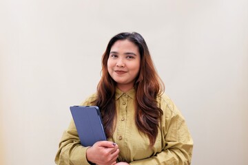 Fototapeta premium Warm smile radiates from a confident woman holding a clipboard. Her hands grip the folder, poised for work or presentation. Soft beige backdrop highlights her calm, professional demeanor.