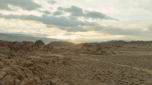 A cinematic landscape shot of the Alabama Hills in California reveals massive desert rock formations with snow-covered Sierra Nevada mountain peaks rising in the background beneath dramatic clouds. Th