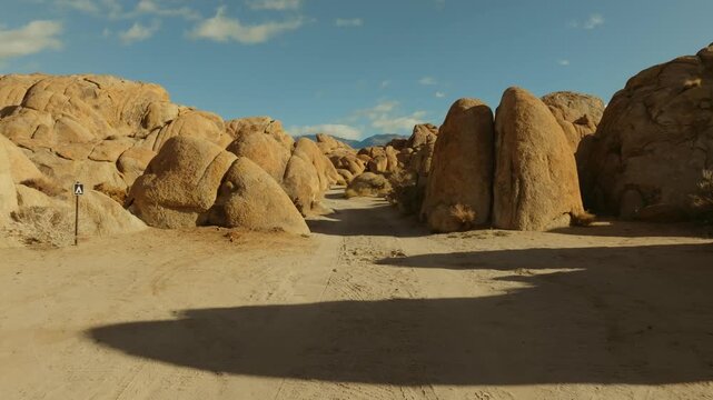 A cinematic landscape shot of the Alabama Hills in California reveals massive desert rock formations with snow-covered Sierra Nevada mountain peaks rising in the background beneath dramatic clouds. Th
