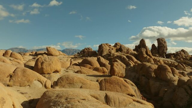 A cinematic landscape shot of the Alabama Hills in California reveals massive desert rock formations with snow-covered Sierra Nevada mountain peaks rising in the background beneath dramatic clouds. Th