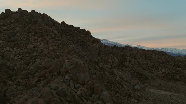 A cinematic landscape shot of the Alabama Hills in California reveals massive desert rock formations with snow-covered Sierra Nevada mountain peaks rising in the background beneath dramatic clouds. Th