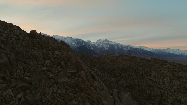 A cinematic landscape shot of the Alabama Hills in California reveals massive desert rock formations with snow-covered Sierra Nevada mountain peaks rising in the background beneath dramatic clouds. Th