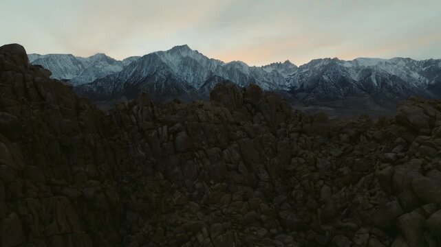 A cinematic landscape shot of the Alabama Hills in California reveals massive desert rock formations with snow-covered Sierra Nevada mountain peaks rising in the background beneath dramatic clouds. Th