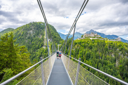Visitors walk on Highline 179, a suspension bridge in Austria. The bridge connects an old castle ruin with a Roman fortress across a wide river gorge surrounded by mountains and trees.
