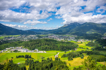 Fototapeta premium A view captures the district town of Reutte in Tyrol, Austria, with green fields, trees, and mountains. Many clouds drift across a blue sky. It is a sunny day with bright sunlight.