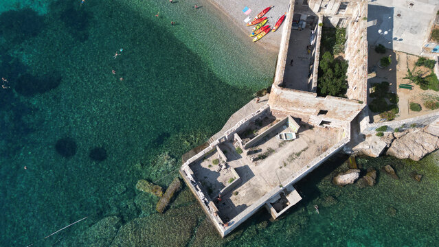 Aerial view of historic stone ruins by the sea with a pebble beach, colorful kayaks, and people swimming in clear turquoise water in Komiza, Split-Dalmatia County, Croatia.