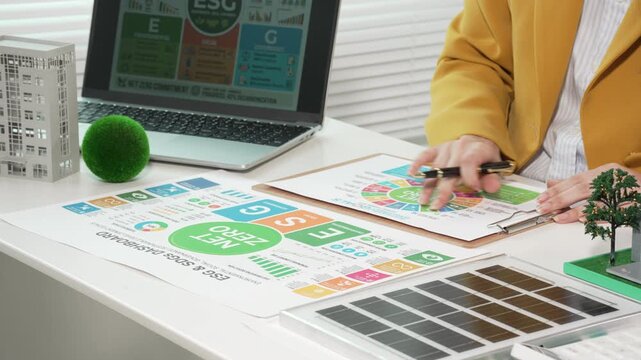Businesswoman reviewing ESG sustainability report at desk with laptop, charts, green icons, building model and mini trees, concept of environmental performance and corporate responsibility.