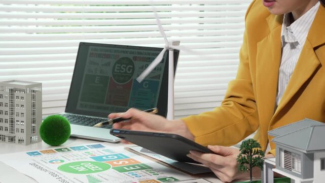 Businesswoman reviews ESG energy efficiency report at desk with laptop, tablet and documents, wind turbine model and building miniatures, bright office light.