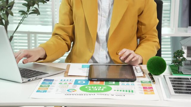 Businessperson in yellow blazer working at desk with laptop and tablet, reviewing analytics charts and eco or renewable energy report; modern office with small model house and green plant decor.