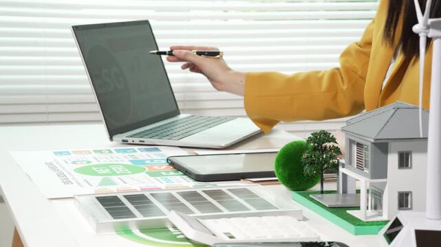 Designer reviewing sustainable home energy plans at desk with laptop, tablet, solar panel samples, green model house and wind turbine, eco friendly architecture concept
