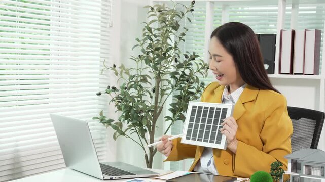 Smiling businesswoman in yellow blazer shows solar panel sample while using laptop at bright office desk with blinds and plants, promoting renewable energy and sustainability.