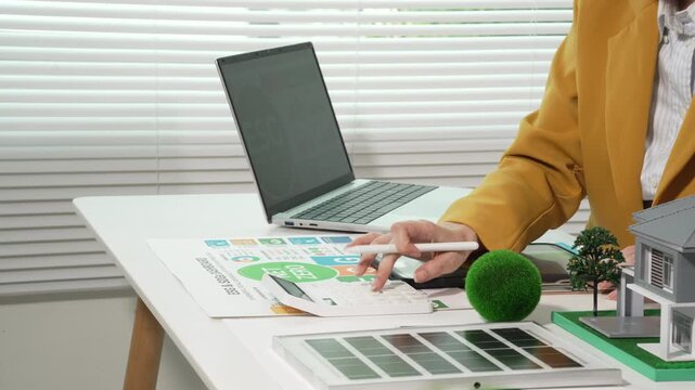 Consultant or architect presenting sustainable housing plan with laptop, model house, solar panels samples, and charts on desk in modern office.