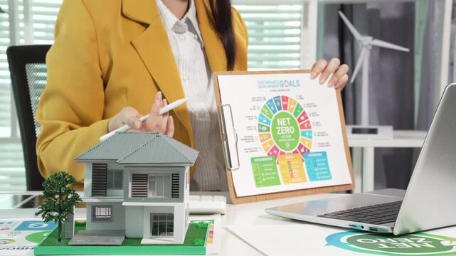 Business professional presenting net zero goals chart beside eco house model and laptop in modern office, sustainability planning and green energy concept.
