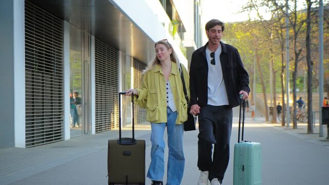A loving couple strolls side by side, grasping hands as they pull their luggage down the busy sidewalk, creating an intimate moment filled with excitement and anticipation for their journey ahead.