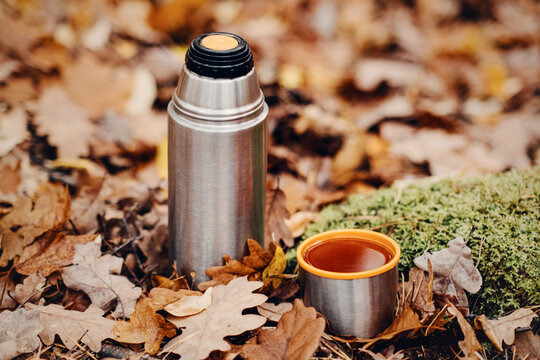Metal thermos and cup filled with tea stand on ground covered in autumn leaves and moss. plastic free lifestyle, conscious consumption habits, durable camping accessories.
