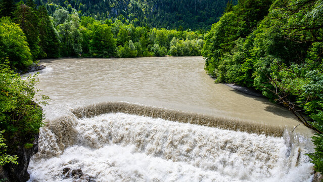 Water from the Lech Falls drops almost 8 meters into the gorge below. Surrounding trees and rocks form a natural setting. The scene shows the force of nature and flowing water.
