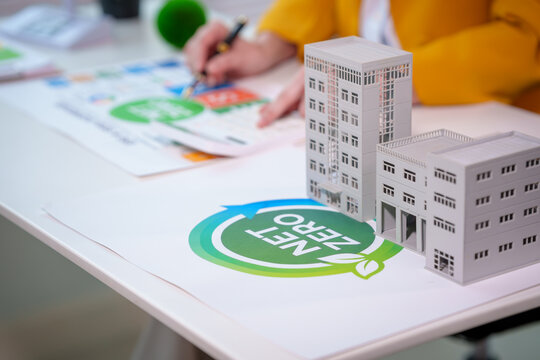 Close-up of sustainable real estate planning with building models on desk and net zero energy symbol, person drafting eco design in office.