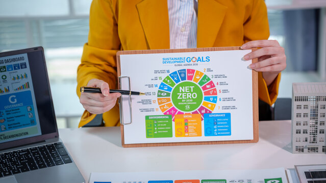Business professional presenting net zero and sdg sustainability goals on clipboard at office desk with laptop and model building, corporate esg strategy meeting concept.