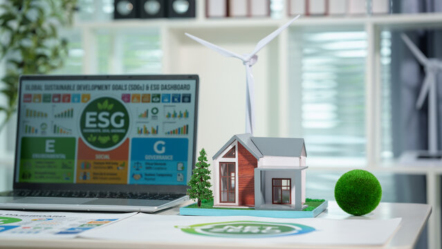 Esg sustainability concept on office desk with laptop dashboard, miniature eco house model, small wind turbine, and greenery in modern workspace.