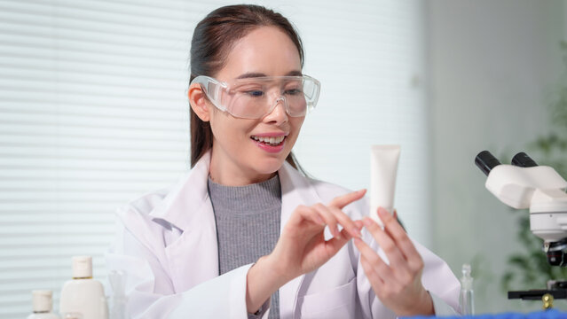 Female scientist in lab coat and safety goggles examining cosmetic product tube in laboratory, microscope on desk, bright modern research setting.