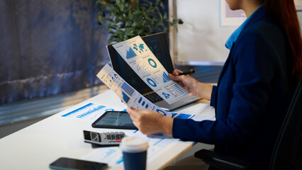 Businesswoman in office reviewing printed analytics reports with charts and graphs, using tablet...