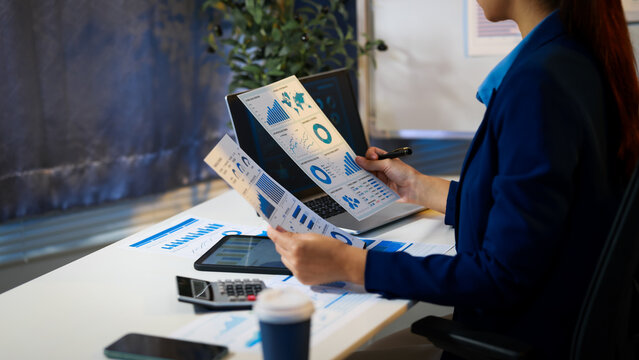 Businesswoman in office reviewing printed analytics reports with charts and graphs, using tablet and laptop on desk with calculator and coffee.