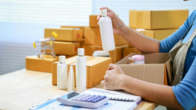 Small business owner packing skincare bottles into shipping boxes at home, with calculator and paperwork on table, e-commerce fulfillment and inventory concept.