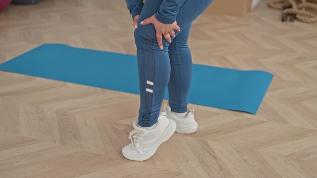 Woman hands on calf above blue yoga mat in studio performing a gentle standing leg stretch; calm recovery.