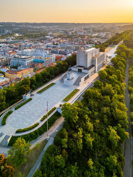 Sunrise lights up the National Monument at Vitkov in Prague. The monument features the Tomb of the Unknown Soldier surrounded by trees and city buildings.