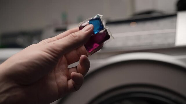 Housewife hand placing colorful laundry detergent pod capsule inside washing machine drum filled with dirty clothes, getting ready to start wash cycle for household cleaning. Shooting in slow motion.