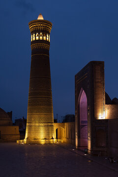 Kalyan Minaret and Kalyan Madrasah illuminated at night on Poi Kalyan Square in Bukhara. Dramatic golden and purple lighting highlights ornamental brickwork against a deep blue twilight sky.