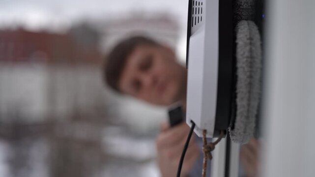 Young man controlling modern robotic window cleaner with remote from inside apartment, ensuring exterior glass spotless during cold winter day with snow outside. Shooting in slow motion.