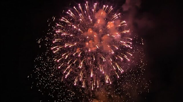 Vibrant golden and pink fireworks exploding in the dark night sky with shimmering sparks and smoke trails. High-altitude pyrotechnic display during a festive outdoor celebration.