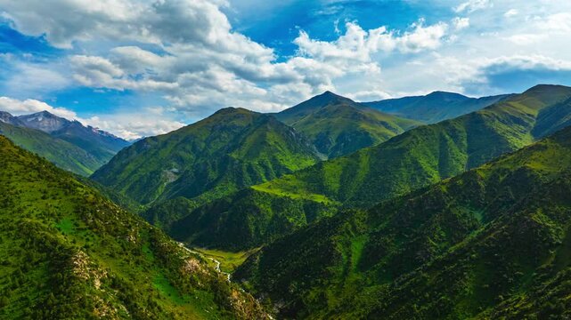Static view of green valley with forest patches and distant peaks. Fast clouds pass overhead, shifting light and shadow across mountainous terrain.