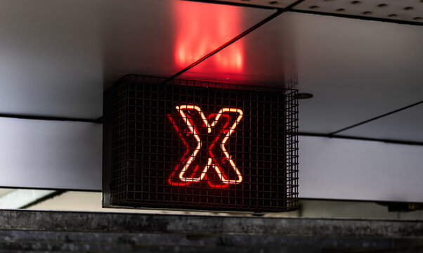Night photo of an illuminated red neon X symbol inside a metal security cage, signalling a restricted entry point or closed lane in a city parking station.
