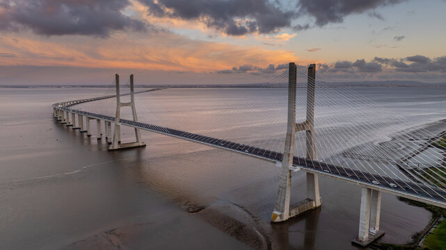 Aerial view of the Vasco da Gama Bridge spanning the Tagus River under a dramatic sunset sky with colorful clouds in Lisboa, Portugal.