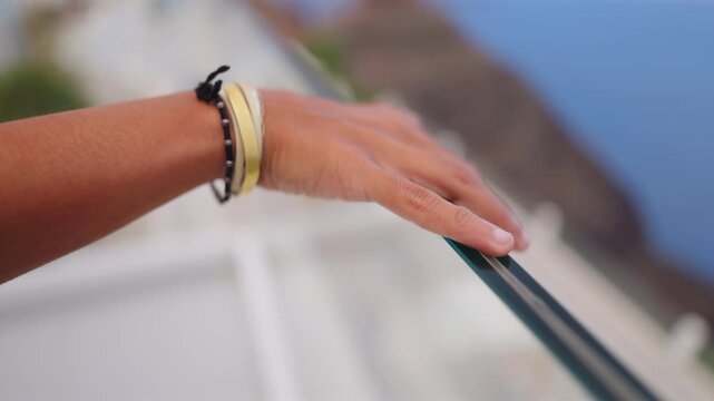 Close-up of woman's hand with gold bangles and black beaded bracelet resting on glass balcony railing overlooking blurred white Santorini buildings and blue Aegean Sea on sunny day.