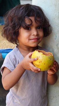 Cute Indian toddler girl drinking fresh wood apple juice directly from its natural shell, happy village child enjoying healthy traditional bel sharbat, authentic rural lifestyle
