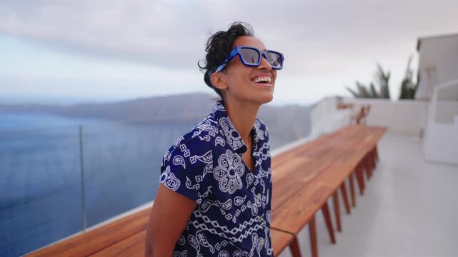 Joyful Indian young woman wearing blue sunglasses and patterned shirt laughs on white rooftop terrace in Santorini Greece. Caldera sea view and mountains create stunning Mediterranean backdrop.