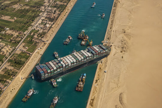 Cargo ship blocking canal, massive container vessel stranded across narrow waterway, aerial drone view of maritime transportation disruption, shipping traffic congestion, commercial freight vessel acc