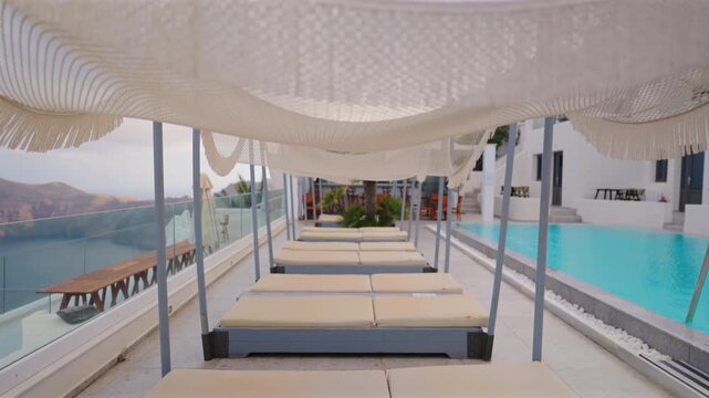 Elegant poolside terrace at luxury resort in Santorini Greece featuring cushioned daybeds under woven canopies with turquoise swimming pool and caldera mountain views at dusk.