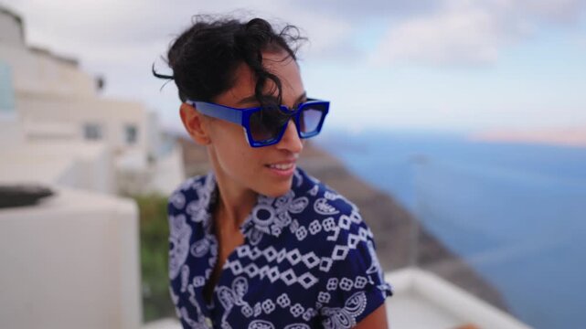 Fashionable young Indian woman wearing blue geometric sunglasses and patterned shirt smiles while enjoying stunning Santorini Greece views with white buildings and sea backdrop at dusk.