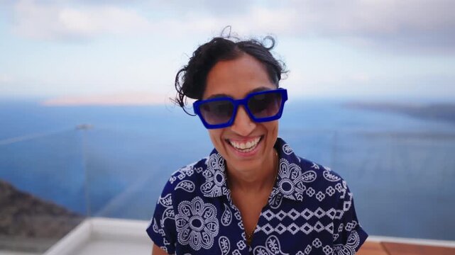 Joyful woman wearing blue sunglasses laughs and claps on white terrace overlooking the Aegean Sea in Santorini Greece. Curly hair blows in wind with caldera and cliffs visible in the scenic.