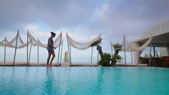 Young Indian woman in summer dress walks barefoot along turquoise infinity pool at luxury resort in Santorini Greece. Decorative canopies and palm trees frame the ocean view under cloudy skies.