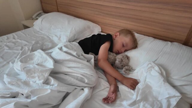 Close-up of a cute little blonde boy sleeping peacefully in his bed while hugging a small plush teddy bear toy.