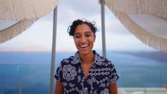 Happy young Indian woman with curly hair laughing genuinely on luxury terrace in Santorini Greece. Decorative fringed umbrellas frame the scene with Aegean Sea and caldera views at dusk.