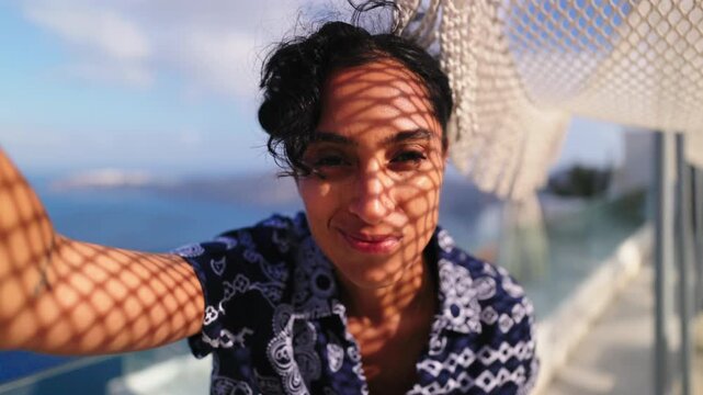 Young woman with curly dark hair takes selfie under woven net umbrella creating diamond shadow patterns on her face. Seaside terrace with blue ocean and islands visible in Santorini Greece.