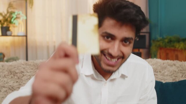 Indian young man at home holding a credit card, looking excited as makes an online purchase. Arabian guy, thrilled, applying the card, anticipating receiving a gift, with look of satisfaction on face